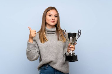 Teenager girl with sweater over isolated blue background holding a trophy clipart