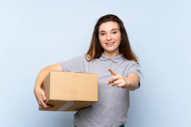 Genç esmer kız izole edilmiş mavi arka planda bir kutu tutuyor ve ön tarafı işaret ederken onu başka bir yere taşıyor.