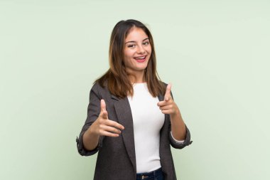 Önü göstererek yeşil arka planda ceketi olan esmer genç kız gülümsüyor.