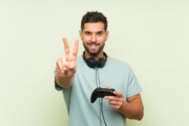Genç yakışıklı adam izole yeşil duvar üzerinde bir video oyunu denetleyicisi ile oynarken gülümseyerek ve zafer işareti gösteren