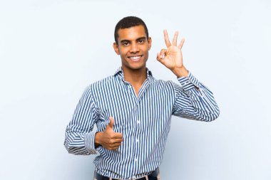 Genç, yakışıklı, esmer, soyutlanmış, mavi arka planda, tamam işareti ve baş parmak hareketi gösteren adam.