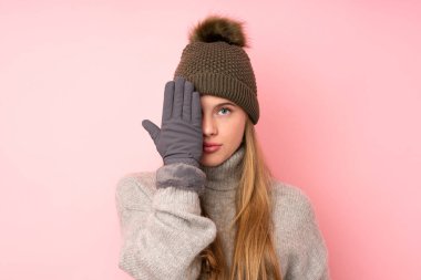 Kışlık şapkalı, pembe arka planı olan genç bir kız. Göz kapağını elleriyle kapatıyor.