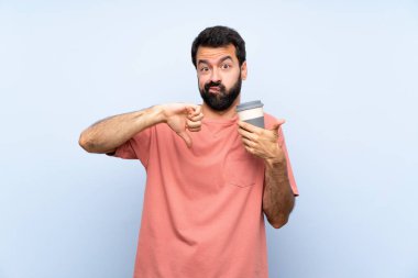 Sakallı genç adam izole edilmiş mavi arka planda kahve tutuyor ve iyiye-kötüye işaret yapıyor. Evet ya da hayır arasında kararsız