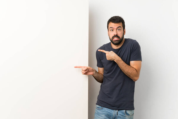 Young handsome man with beard holding a big empty placard frightened and pointing to the side