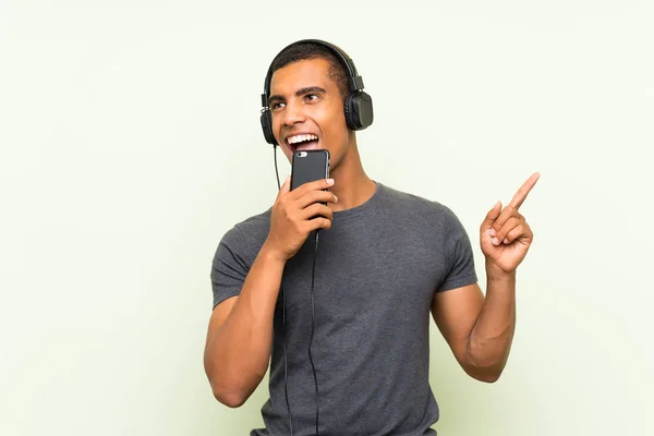 Junger gutaussehender Mann hört Musik mit Handy über isolierte grüne Mauer — Stockfoto