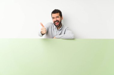 Young handsome man with beard holding a big green empty placard with thumbs up because something good has happened