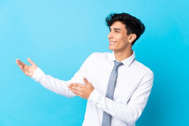 Arjantinli genç iş adamı, izole edilmiş mavi arka planda, davet edilmek için ellerini yana uzatıyor.