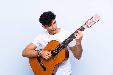 İzole edilmiş mavi arka planda gitar çalan genç Arjantinli adam.