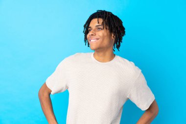 Genç Afro-Amerikalı bir adam soyutlanmış mavi arka planda kolları kalçasında ve gülümsüyor.