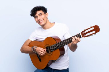 İzole edilmiş mavi arka planda gitar çalan genç Arjantinli adam.