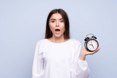 Soyutlanmış mavi arka planda, elinde klasik çalar saat olan genç bir kadın.