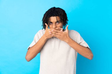 İzole edilmiş mavi arka planda ağzı elleriyle kapatan genç bir Afro-Amerikalı.