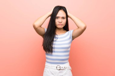 Soyutlanmış pembe arka planda genç Asyalı kız hayal kırıklığına uğradı ve ellerini kafaya koydu.
