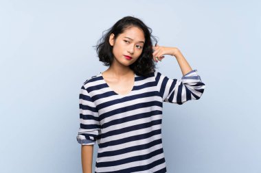 Asyalı genç bir kadın soyutlanmış mavi arka planda deliliğin hareketini yapıyor. Parmağını kafaya koyuyor.