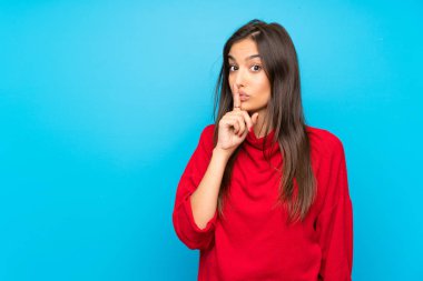 İzole edilmiş mavi arka planda kırmızı kazaklı genç bir kadın sessizlik hareketi yapıyor.