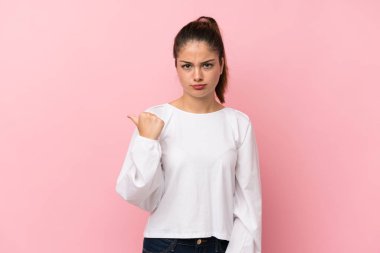 Soyutlanmış pembe arka planda genç esmer kız mutsuz ve kenarı işaret ediyor.