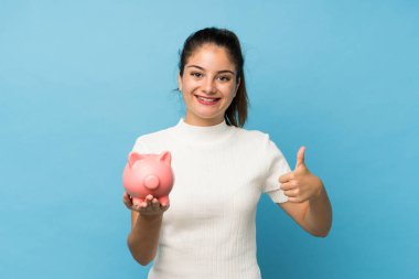 Genç esmer kız izole edilmiş mavi arka planda büyük bir kumbara tutuyor.