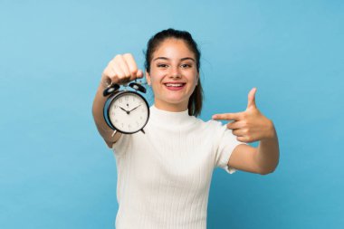 İzole edilmiş mavi arka planda esmer bir kız. Elinde klasik çalar saat var.