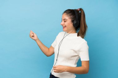 İzole edilmiş mavi arka planda genç esmer kız kulaklık ve dansla cep telefonunu kullanıyor.