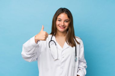 Doktor elbisesi ve başparmağıyla soyutlanmış mavi arka planda genç esmer kız.