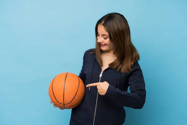 年轻的黑发女孩带着篮球运动越过孤立的蓝色背景 指着篮球运动 — 图库照片