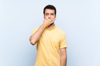 İzole edilmiş mavi arka planda sakallı genç bir adam elleriyle ağzını kapatıyor.