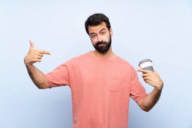 Sakallı genç adam elinde kahve ile soyutlanmış mavi arka planda gururlu ve kendini beğenmiş.