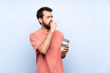 Elinde kahve tutan sakallı genç adam, izole edilmiş mavi arka planda öksürük ve kötü hissetmekten muzdarip.