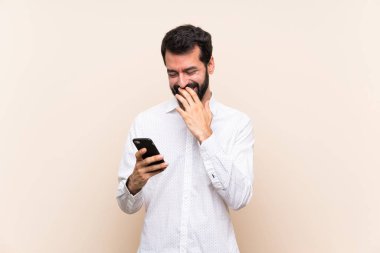 Elinde cep telefonu olan sakallı genç adam çok gülümsüyor.