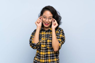 Asyalı genç bir kadın gözlüklü ve şaşırmış bir şekilde mavi arka planda.