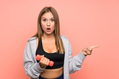 Genç sporcu kız halter kaldırıyor. Pembe arka plan üzerinde. Şaşırmış ve işaret ediyor.