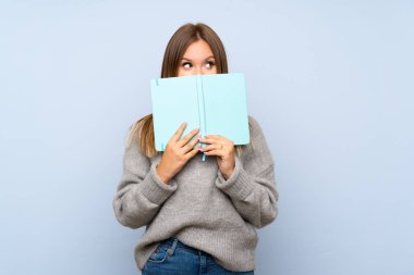 Izole mavi arka plan üzerinde kazak ile Genç kız tutma ve bir kitap okuma