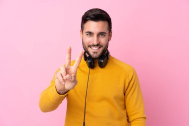 Gözlerden uzak pembe arka planda kulaklıkları olan genç yakışıklı adam gülümsüyor ve zafer işareti gösteriyor.