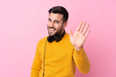 Kulaklıkları olan genç yakışıklı bir adam pembe arka planda mutlu bir ifadeyle selamlıyor.