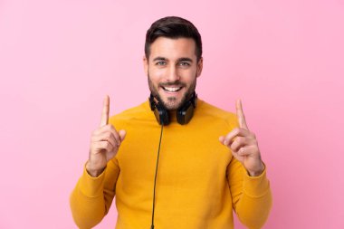 İzole edilmiş pembe arka planda kulaklıkları olan yakışıklı genç adam harika bir fikre işaret ediyor.