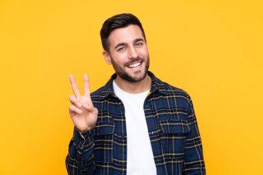 Sarı arka planda sakallı yakışıklı genç adam gülümsüyor ve zafer işareti gösteriyor.