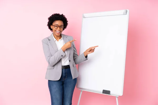 African american business woman giving a presentation on white board over isolated pink background surprised and pointing side