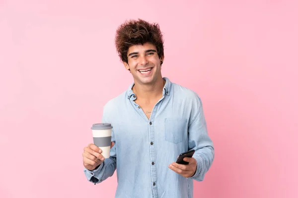 年轻的高加索男子在孤立的粉色背景上拿着咖啡和手机走了 — 图库照片