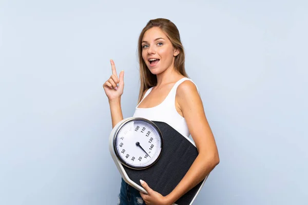 Young blonde girl with weighing machine over isolated blue white — Stock Photo, Image