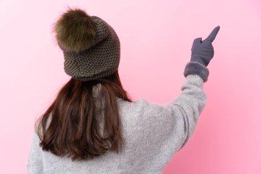Kışlık şapkalı genç Kolombiyalı kız izole edilmiş pembe duvarın arkasında işaret parmağıyla işaret ediyor.