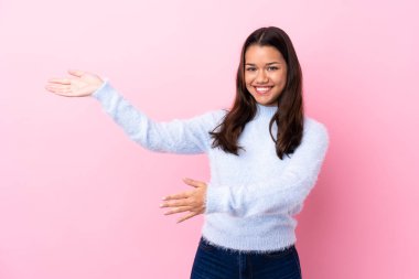İzole edilmiş pembe arka planda genç Kolombiyalı kız. Davet etmek için ellerini yana uzatıyor.