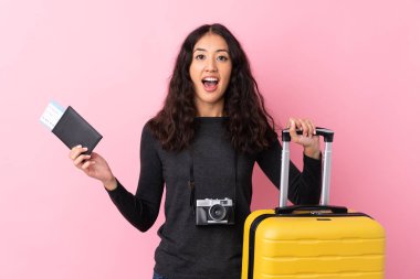 Karmaşık ırk kadını, izole pembe arka planda tatilde, bavullu ve pasaportlu ve şaşırmış.