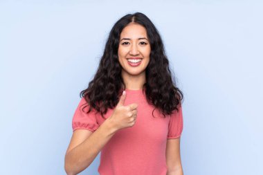 İzole edilmiş mavi arka planda süveter giyen melez bir kadın baş parmak işareti yapıyor.