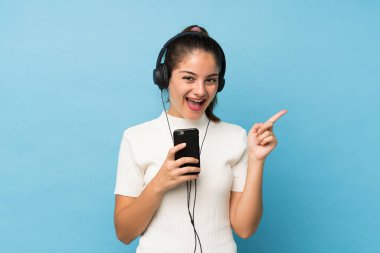 Genç esmer kız mavi arka planda, kulaklıklı ve şarkı söyleyen cep telefonunu kullanıyor.