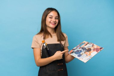 Genç esmer kız izole edilmiş mavi arka planda bir palet tutuyor ve ona doğrultuyor.