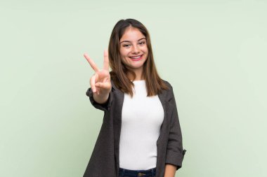 İzole edilmiş yeşil arka planda ceketli esmer kız gülümsüyor ve zafer işareti gösteriyor.