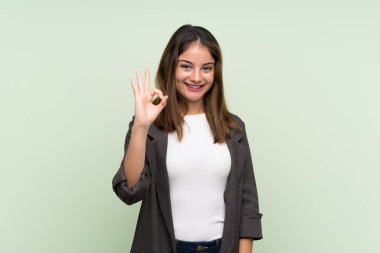 Yeşil arka planda ceketi olan esmer genç kız parmaklarıyla işaret veriyor.