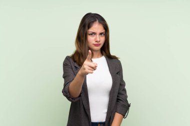 İzole edilmiş yeşil arka planda ceketi olan genç esmer kız hayal kırıklığına uğramış ve ön tarafı işaret ediyor.