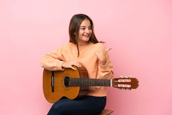 Young brunette girl with guitar over isolated pink background pointing to the side to present a product