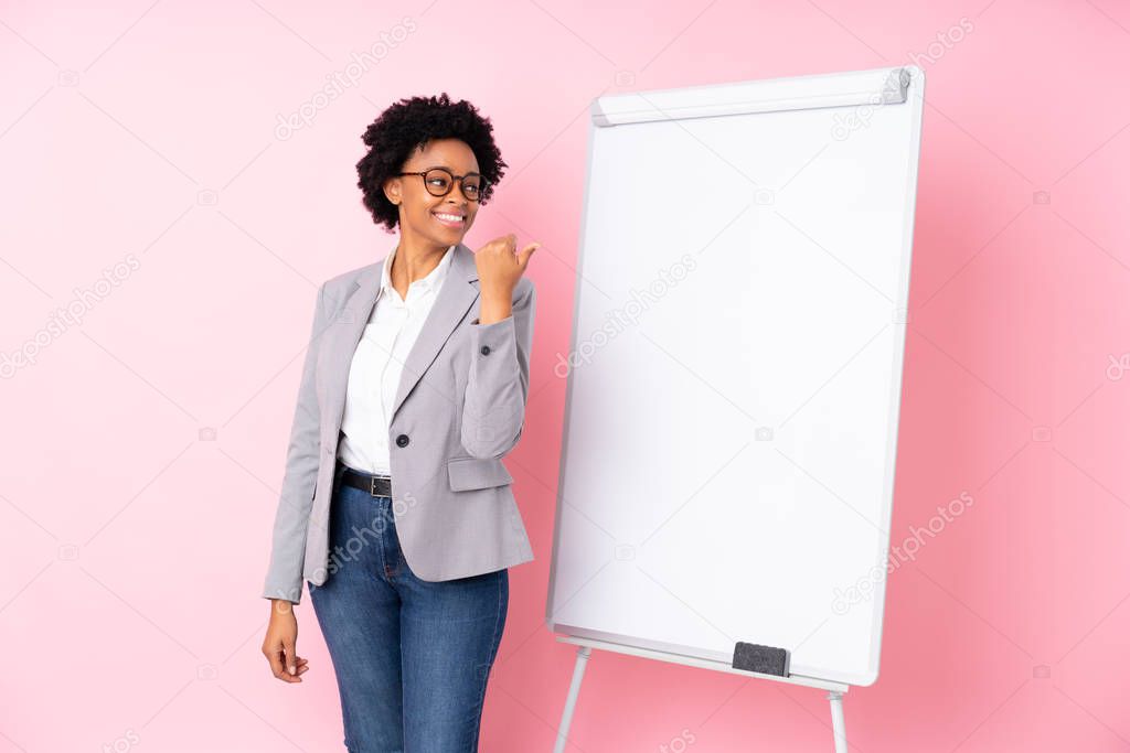 African american business woman giving a presentation on white board over isolated pink background pointing to the side to present a product